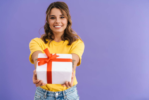 Happy beautiful nice girl smiling and reaching out gift box isolated over purple background - Fotoğraf, Görsel