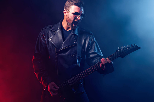 Brutal bearded Heavy metal musician in leather jacket and sunglasses is playing electrical guitar. Shot in a studio on dark background with smoke - Foto, imagen