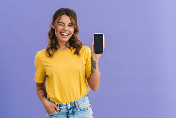 Mujer joven sonriente mostrando teléfono inteligente de pantalla en blanco aislado sobre fondo azul - Foto, imagen