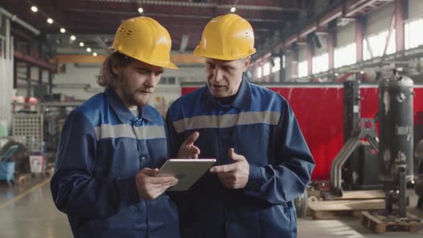 Średnie spowolnienie dwóch robotników fabrycznych w twardych kapeluszach i mundurkach omawiających pracę w zakładzie, patrzących na cyfrowy tablet, a następnie obracających głowice do kamery - Materiał filmowy, wideo