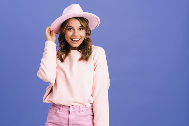 Happy charming girl in hat smiling and looking at camera isolated over purple background - Fotó, kép