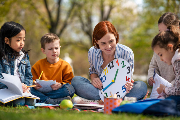 Lehrer mit kleinen Kindern sitzen draußen im Stadtpark, Lerngruppenpädagogik. - Foto, Bild