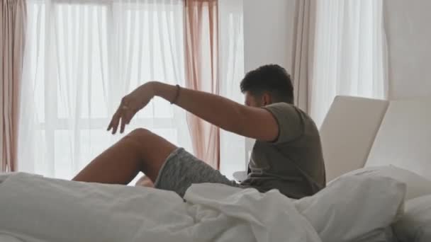Vista trasera de mediana toma de hombre joven de etnia latina levantándose por la mañana y estirándose en una cama acogedora mirando a la ventana panorámica - Metraje, vídeo