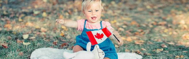 Очаровательная маленькая белая малышка, размахивающая канадским флагом в парке на улице. Ребенок гражданин, сидящий на земле в парке и празднующий День Канады 1 июля. Заголовок веб-баннера. - Фото, изображение