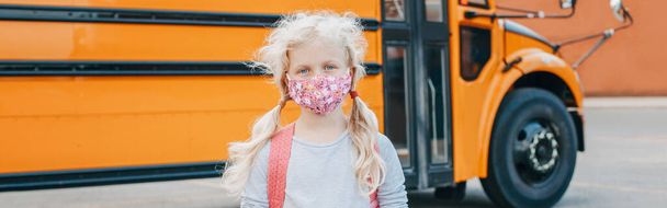 Μια μαθήτρια με μάσκα προσώπου που στέκεται δίπλα σε ένα κίτρινο λεωφορείο. Παιδί με μέσα ατομικής προστασίας στο πρόσωπο. Εκπαίδευση και πίσω στο σχολείο. Νέο φυσιολογικό κατά τη διάρκεια του κορωνοϊού. Τίτλος banner ιστού. - Φωτογραφία, εικόνα