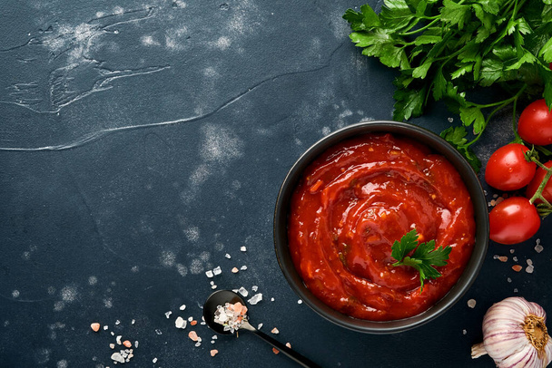 Tomato ketchup spicy sauce in a black bowl with spices and fresh tomatoes on a dark slate, stone or concrete background. Top view with copy space. - Foto, Bild