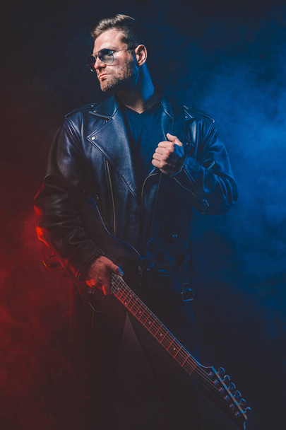 Brutal bearded Heavy metal musician in leather jacket, cap and sunglasses is playing electrical guitar. Shot in a studio on dark background - Photo, Image