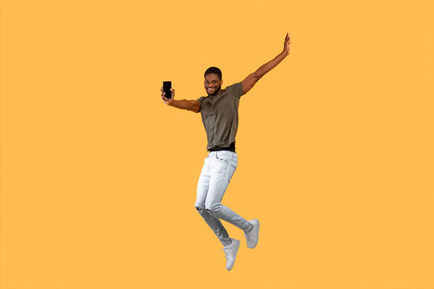 Radosny Afrykański Amerykanin skacze po żółtym tle i pokazuje smartfon z czarnym ekranem do kamery. - Zdjęcie, obraz