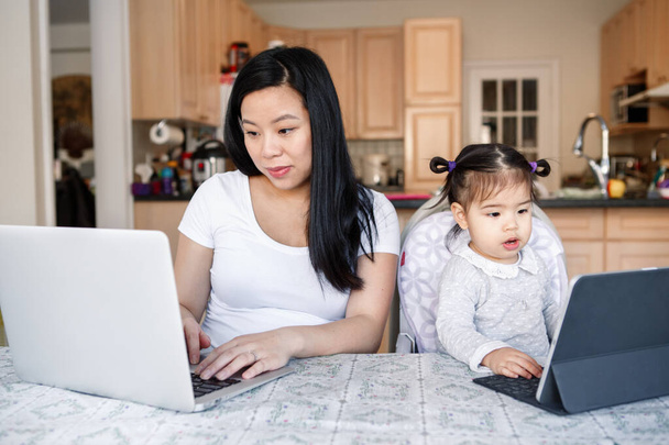 Aziatische Chinese moeder met dochter baby werken samen op laptop, tablet. Werkplaats van freelancer vrouw met kind. Blijf thuis mam werkt op afstand baan. Online onderwijs thuisonderwijs. Nieuw normaal - Foto, afbeelding