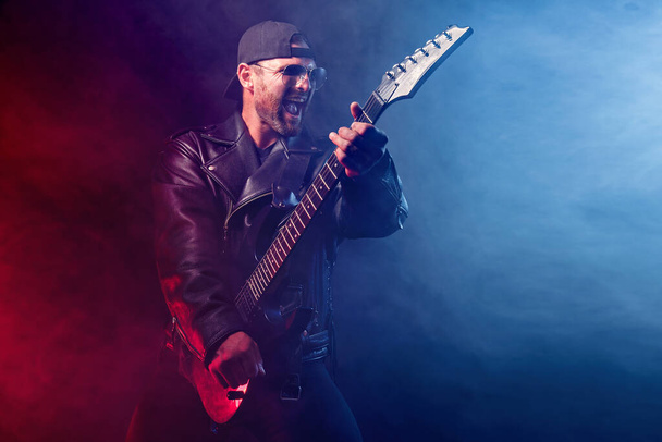 Brutal bearded Heavy metal musician in leather jacket and sunglasses is playing electrical guitar. Shot in a studio on dark background with smoke - Foto, imagen