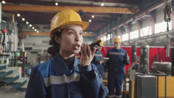 Slowmo close-up of young female plant supervisor in hard hat and uniform giving instructions on walkie-talkie then looking at camera while male colleagues standing in background - Footage, Video