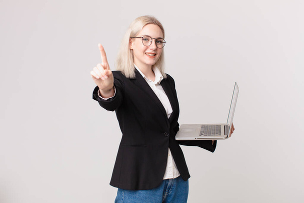 blond pretty woman smiling proudly and confidently making number one and holding a laptop - Foto, Bild