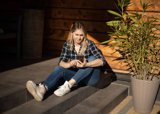 Młoda kobieta siedzi na schodach na ganku w letni dzień i za pomocą smartfona do nauki online - Zdjęcie, obraz