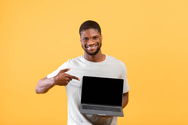 Leuke website. Positieve Afro-Amerikaanse man wijst naar laptop met zwart leeg scherm, kopieer ruimte voor uw ontwerp - Foto, afbeelding