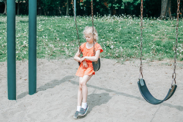 Trauriges unglückliches Mädchen schaukelt allein auf Schaukel im Park. Aufgebrachte gelangweilte Kinder warten auf dem Spielplatz auf ihren Freund. Einsames Kind schaukelt allein im Park und vermisst Freund. Negative Emotionen. - Foto, Bild