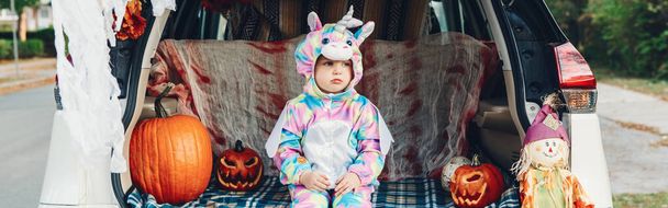 Karkki tai takakontti. Surullinen järkyttynyt vauva yksisarvinen puku juhlii Halloween auton takakontissa. Söpö taapero juhlii lokakuun loma ulkona. Turvallinen vaihtoehtoinen juhla. Web-bannerin otsikko. - Valokuva, kuva