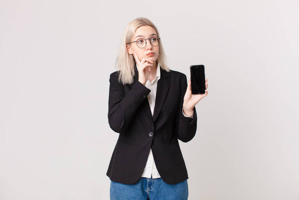 blond pretty woman thinking, feeling doubtful and confused and holding a mobile telephone - Photo, image