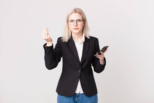 blond pretty woman making capice or money gesture, telling you to pay and holding a mobile telephone - Fotoğraf, Görsel