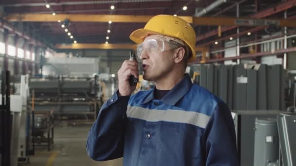 Vakavan aikuisen tehtaan esimiehen keskikokoinen hitaus turvalaseissa, kypärässä ja univormussa antamassa ohjeita radiopuhelinta käyttäville työntekijöille - Materiaali, video