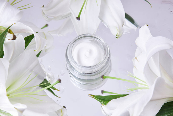 Cream for skin care, natural cosmetics made of flowers and petals. A glass jar of white cream stands among the lily flowers - Photo, image