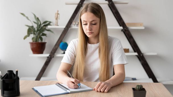 A estudante caucasiana concentrada sentada em uma mesa no local de trabalho é treinada escrevendo informações em um caderno. Um professor ou tutor se prepara para uma lição. - Foto, Imagem