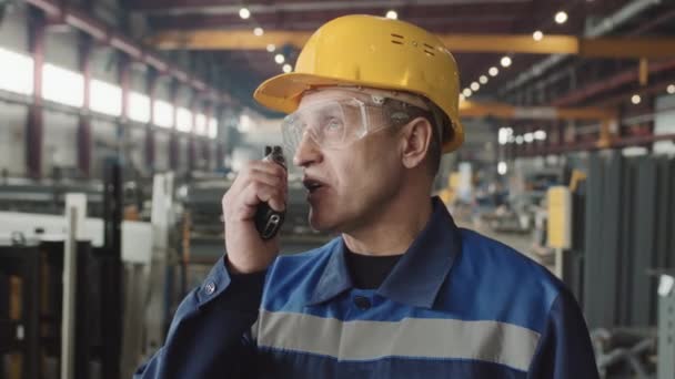 Retrato de cerca del supervisor de planta senior en gafas de seguridad y sombrero duro hablando en walkie-talkie y luego mirando a la cámara durante el turno - Imágenes, Vídeo