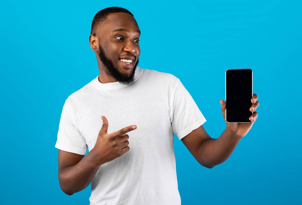 Czarny facet wskazując palec na smartfon pusty ekran, niebieskie tło - Zdjęcie, obraz