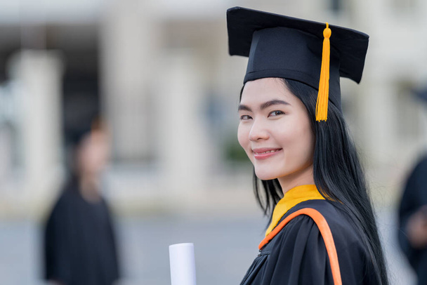 A young beautiful Asian woman university graduate in graduation gown and mortarboard holds a degree certificate stands in front of the university building after participating in college commencement - Photo, image