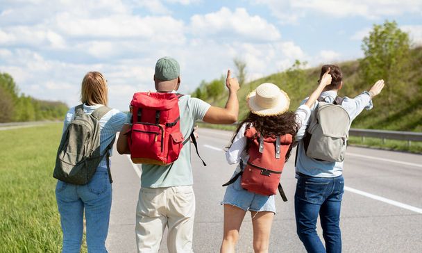Back view of young multiracial friends standing on highway, showing hitchhiking gesture, waving down car outdoors - Foto, Imagen