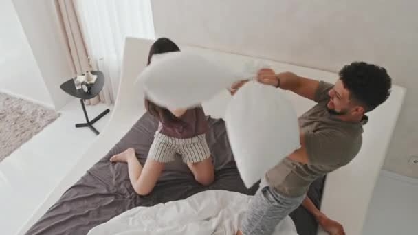 Vista superior de la joven pareja latina juguetona teniendo pelea de almohadas en la cama disfrutando de la mañana juntos en casa en su moderno apartamento de estilo minimalista - Metraje, vídeo