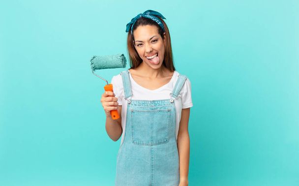 young hispanic woman with cheerful and rebellious attitude, joking and sticking tongue out and painting a wall - Foto, afbeelding