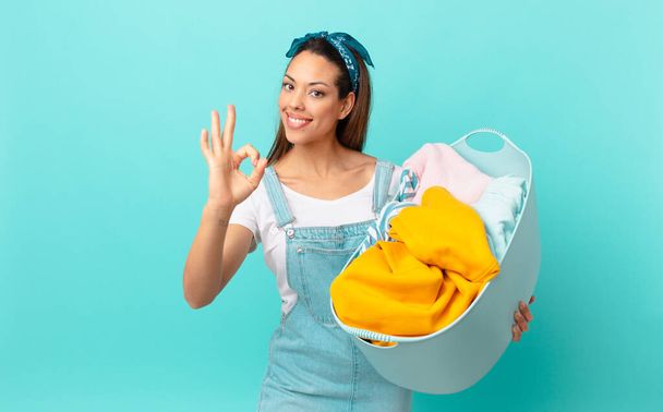 young hispanic woman feeling happy, showing approval with okay gesture and washing clothes - 写真・画像