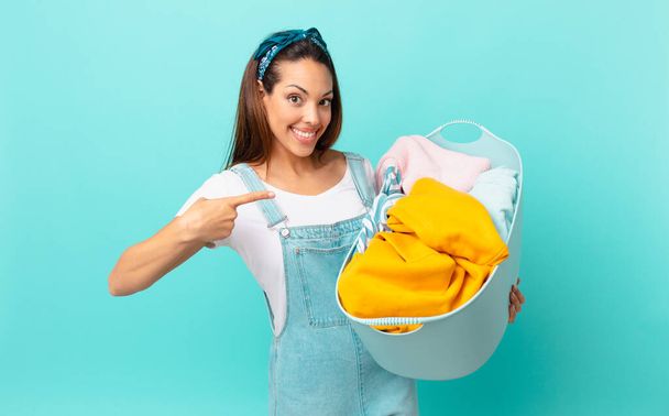 young hispanic woman smiling cheerfully, feeling happy and pointing to the side and washing clothes - Foto, Imagen