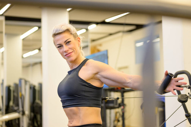 Lächelnde, freundliche, athletische Frau, die in einem Fitnessstudio an Widerstandsgeräten trainiert, um ihre Kernmuskulatur in einem Gesundheits- und Fitnesskonzept zu stärken und zu straffen - Foto, Bild