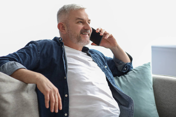 Χαλαρός ώριμος άντρας κάθεται στον καναπέ και μιλάει στο τηλέφωνο. - Φωτογραφία, εικόνα
