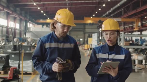 Medium slowmo of couple of professional factory workers discussing project plan looking at tablet while walking through plant facility wearing safety glasses and hard hats - Footage, Video