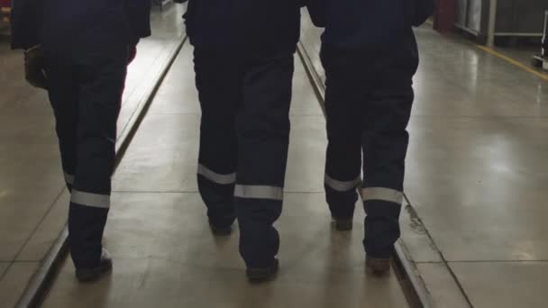 Umgeknickte Rückansicht mit langsamem Tempo von drei männlichen und weiblichen Arbeitern in Overall-Uniformen und harten Hüten, die während der Schicht durch das Werk laufen - Filmmaterial, Video