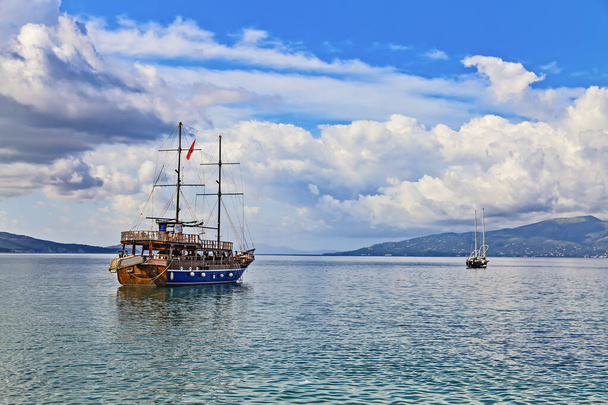 黒の双頭ワシと赤の状態アルバニアの旗を持つアルバニアのサランダ湾での海のツアーのためのヴィンテージマスト木製セーリング船。水平線に浮かぶ小さなヨットとコルフ島 - 写真・画像