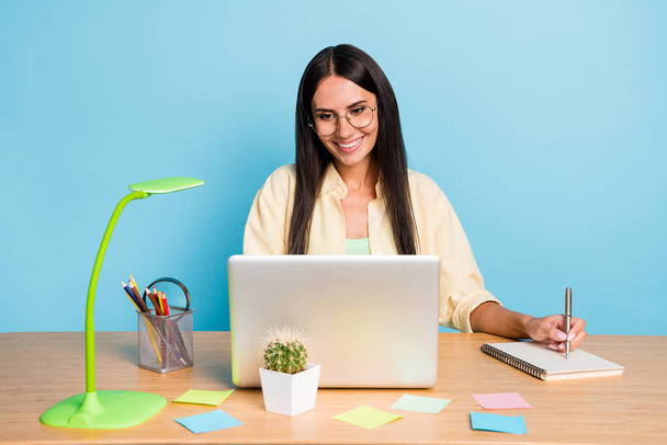 明るい幸せな若いブルネットの女性の写真はノートパソコンの座って机の仕事のノートはパステルブルーの色の背景に隔離された - 写真・画像