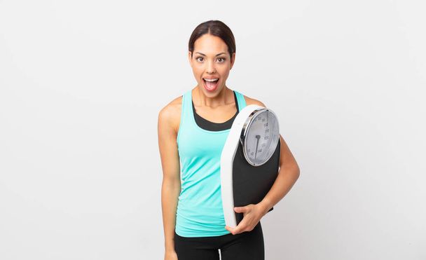 young hispanic woman looking very shocked or surprised and holding a scale - Fotó, kép