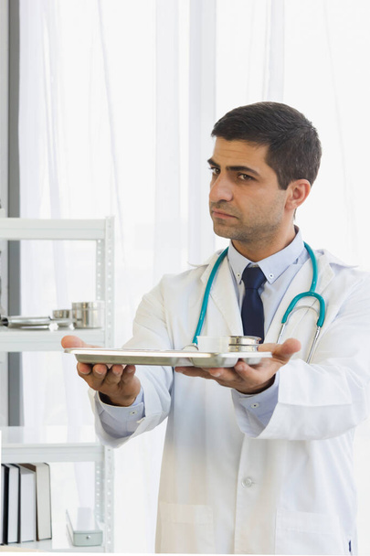Arzt hängte Stethoskop an, hob Medikamententablett mit beiden Händen, hielt sich im Krankenhauszimmer nach vorne - Foto, Bild