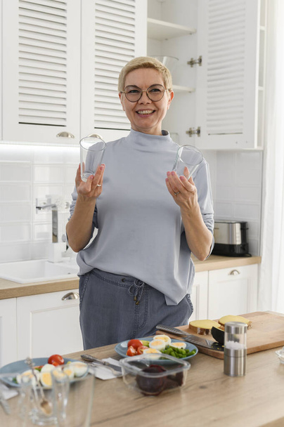 Mulher sênior sorridente faz smoothie saudável com legumes na cozinha. Segura copos vazios, oferece bebida - Foto, Imagem