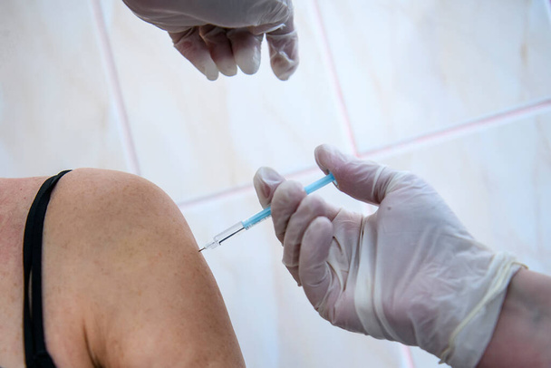Закрыть руки работнику здравоохранения, который вакцинирует пожилого человека коронавирусом. Руки в защитных перчатках держат шприц с вакциной. Высокое качество фото - Фото, изображение