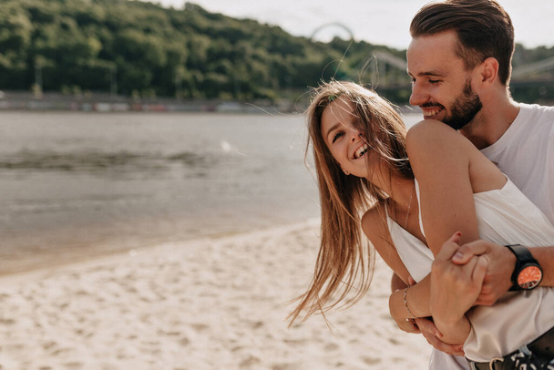 Χαμογελαστό χαρούμενο κορίτσι με μακριά ιπτάμενα μαλλιά αγκαλιάζονται με το αγόρι της στην παραλία κοντά στο ποτάμι στο φως του ήλιου. Υψηλής ποιότητας φωτογραφία - Φωτογραφία, εικόνα