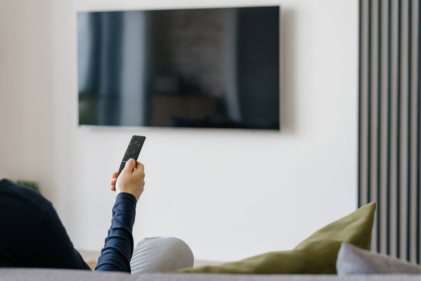 Modern living room interior with blank tv screen panel on blurred white wall background. Cropped shot of woman with remote control in hand sitting on sofa in room with minimalist functional design - Zdjęcie, obraz