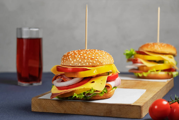Two burgers on a wooden board with bacon, pepper, tomato, lettuce, cucumber, cheese. The second burger in blur. Grey background. Wooden skewers. - Photo, image