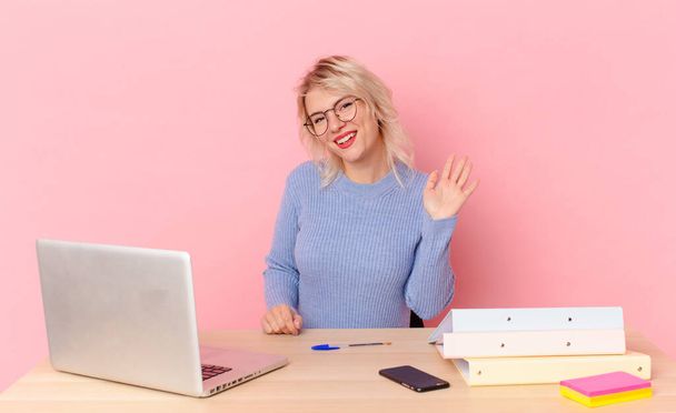 blonde pretty woman young pretty woman smiling happily, waving hand, welcoming and greeting you. workspace desk concept - Fotoğraf, Görsel
