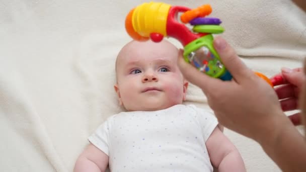 Jonge moeder schudden en spelen met kleurrijke plastic speelgoed met haar kleine baby liggend in wieg. - Video