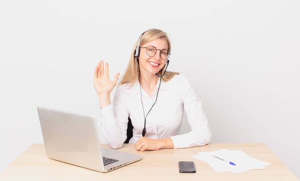 blonde pretty woman young blonde woman smiling happily, waving hand, welcoming and greeting you and working with a laptop - Фото, изображение