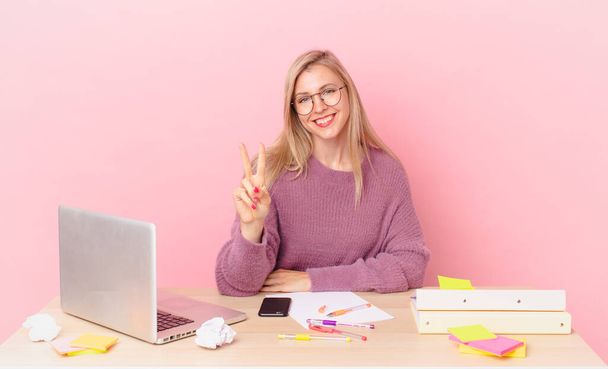 blonde pretty woman young blonde woman smiling and looking happy, gesturing victory or peace and working with a laptop - Фото, изображение